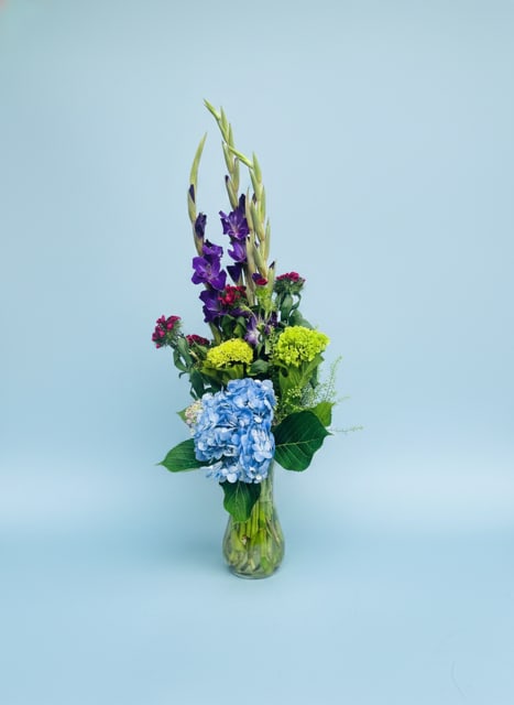 Gladiolas-Hydrangea Bouquet - Tall, airy stems and a bold blue hydrangea give this upright flower arrangement an immediate, modern presence-perfect as a birthday bouquet, a Thinking of You gift, or a Father's Day surprise that feels fresh and confident. The vertical silhouette sends a bright, uplifting message that works equally well for Get Well wishes, Housewarming, or a gentle I'm Sorry gesture.  Blue hydrangea anchors the base while purple gladiolus blooms rise through the center, complemented by soft pink clusters, creamy gardenias, and cheerful green accents for contrast and texture. Arranged in a clear glass vase, the composition reads contemporary and light, with a lively mix of colors-blue, green, yellow, purple, and pink-that catch the eye without overwhelming.  This arrangement suits many moments: a new baby celebration, sympathy or funeral when a refined, upright piece is preferred, or simply to congratulate someone. Vase-ready and designed to make the recipient feel noticed, it arrives ready to display; for best longevity, keep in fresh water and out of direct heat.