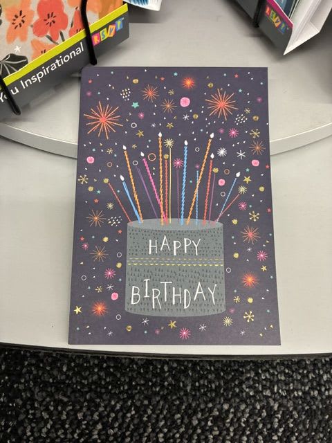 Happy Birthday Card - A dark purple birthday card that feels like a tiny celebration in your hands - the central gray cake illustration is surrounded by playful, sparkler-like bursts and starbursts in bright yellow, pink, blue and black accents. The hand-drawn lettering and whimsical, casual style make this greeting card a cheerful complement to a birthday bouquet or a Thinking of You surprise, instantly setting a festive, personal tone for the recipient.  Printed on sturdy card stock with a matching envelope, the design's mixed-color palette pops against the deep background and works well paired with colorful arrangements or as part of a gift set. Size and exact paper weight are standard for boxed greeting cards; include a short handwritten message inside to make the moment feel truly seen - perfect for birthdays, small celebrations, or to brighten someone's day any time of year.