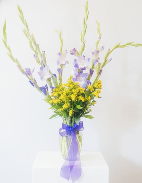 Happy Gladiolus - Lift their spirits with tall, airy gladiolus and delicate lavender-and-white blooms that rise above a sunny cluster of bright yellow filler flowers-this flower arrangement is a cheerful birthday bouquet and a perfect just-because or thank-you gift. The loose, vertical shape reads fresh and springlike, making it equally fitting for a housewarming, new baby welcome, retirement celebration, or to brighten a table at a wedding or Father's Day brunch. Recipients will notice the graceful height and whisper-soft color contrast first, and feel genuinely seen by the light, whimsical presentation.  Arranged in a clear glass vase and finished with a sheer purple ribbon, the design pairs soft purples and whites with vivid yellow and green accents for lively seasonal appeal without feeling tied to one holiday. The composition's airy stems and compact yellow center create a pleasing textural contrast that looks hand-tied yet gift-ready; it works well for spring, Easter, Rosh Hashanah, Mother's Day, Best Friends Day, or as a New Year pick-me-up. Simple care-fresh water in the vase and a cool spot away from direct sun-keeps the arrangement looking its best, and the vertical size makes it an elegant choice for tabletops, entryways, or bedside displays.