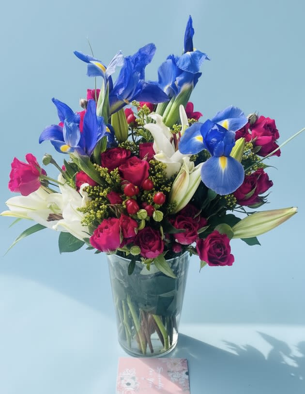 July Vibes - Vivid blue irises paired with bright pink roses and crisp white lilies create a lively, celebratory statement-perfect as a summer birthday bouquet or a Just Because surprise. This flower arrangement for occasions like Father's Day, Thank You, Anniversary, or Thinking of You brings an energetic mix of red, white, blue, green, and pink tones that read both festive and polished, so it also suits Fourth of July or Memorial Day gatherings without feeling holiday-limited.  Densely packed in a clear glass vase, the rounded upright shape highlights the contrast between cool blue iris petals and the warm, velvety roses, while the white lilies add height and a clean, fragrant touch. The compact design makes it easy to display on a dining table, desk, or bedside table; its fresh, tightly arranged composition keeps a neat silhouette that reads as thoughtfully arranged rather than casual.  Choose this arrangement to celebrate a new baby, send congratulations, or make someone feel genuinely seen-she'll feel brightened by the color and scent. Care is simple: trim stems and refresh water every few days to extend vase life; available in the pictured size for immediate gifting.