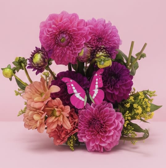 Midsummer's Butterfly Dream - A cheerful cluster of large pink and purple dahlias takes center stage in this whimsical birthday and spring bouquet, accented by peach blooms and sunny yellow filler that brighten the composition. The decorative pink butterfly perched near the center gives the arrangement a playful, romantic feel-perfect for saying "thinking of you," celebrating a quinceañera, or surprising someone just because they deserve to smile.  Soft pastels and textural buds create a compact, hand-tied look that reads feminine and fresh without being fussy. Mixed carnations and other complementary blooms tuck between the dahlias for fullness and lasting color; the yellow accents add light and lift at the edges. This arrangement is sized for tabletop display and arrives ready to place in a vase-an inviting, easy way to make someone feel truly seen on birthdays, romantic moments, or simple springtime celebrations.