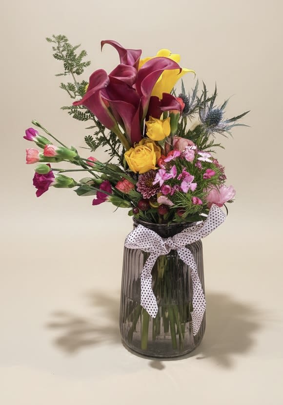 Mini gift jar medley - Vivid magenta and sunny yellow calla lilies make the first impression in this Mini gift jar medley, a cheerful flower arrangement perfect for a birthday, Get Well wish, or a thoughtful Just Because surprise. The asymmetrical, airy design gives the recipient a sense of joyful movement-the kind of bouquet that reads as both celebratory and intimately personal, the sort of gift that says you're thinking of them.  Smaller spray roses, peonies, carnations and mini calla lilies in pink, coral, white and purple tuck around the taller stems to create a lively mixed bouquet with layered texture and color. The flowers arrive gathered in a dark ribbed glass vase and finished with custom ribbon for a dressed-up yet casual look that suits springtime celebrations or everyday brightening.  Ideal for someone recovering, celebrating a milestone, or simply needing a mood lift, this compact arrangement fits on a bedside table, desk, or kitchen counter and brings instant cheer. Because it's arranged in a water-holding jar, it's ready to enjoy on arrival-refresh the water every few days and trim stems slightly to keep blooms looking fresh longer.