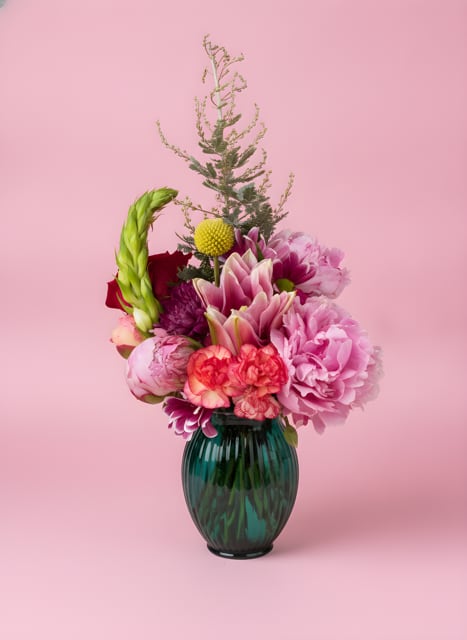Mystery Mini bouquet - A lively cluster of mixed roses, lilies, carnations, and chrysanthemums in a compact, sculptural design-this Mystery Mini bouquet brings bright, varied color and texture to any Just Because, Birthday, or Thinking of You moment. The arrangement reads romantic and cheerful with its mix of soft pinks, coral and white balanced against deeper reds, purples and pops of yellow and orange, making it equally right for Love and Romance, Mother's Day, or a summer celebration like Father's Day when you want something unexpectedly fresh.  Small but expressive, each bouquet is gathered tight at the base with airy stems rising for height and movement, and is presented in your choice of a clear glass vase, a deep green glass vase, or a black textured vase for a modern contrast. Expect layered textures from full-headed roses and chrysanthemums to the softer lilies and sprightly greenery; sizes are compact for tabletops and desks, perfect for brightening a nursery, office, or bedside table for a new baby or new home. Order as a cheerful single surprise or pair for a coordinated gift set-care is simple: fresh water and a cool spot keep blooms looking their best for several days.