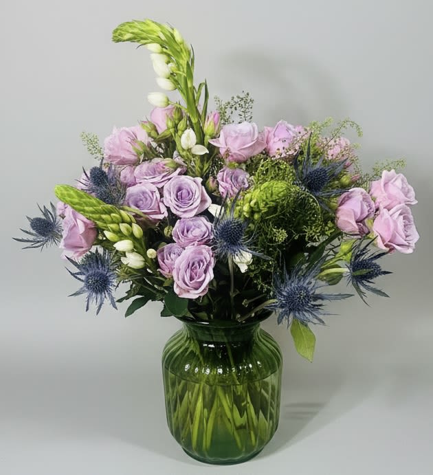 Mythology Bouquet - Lavender roses clustered in a soft, rounded design give this flower arrangement a dreamy, romantic first impression-perfect as a birthday bouquet, a "thinking of you" surprise, or a just-because gift that makes someone feel truly seen. Spiky blue thistle-like blooms and a few tall white buds weave playful contrast into the pastel palette of purple, green, blue, white, and pink, so it also reads beautifully for housewarming, new baby celebrations, or wedding and anniversary moments.  Arranged in a substantial vintage glass vase, the Mythology Bouquet balances delicate lisianthus and mixed blooms with more structured accents for texture and movement. The vase's rich color grounds the arrangement and doubles as a keepsake, so the recipient can enjoy flowers now and use the vessel later. Ideal for occasions from Father's Day and Mother's Day to thank-you gestures or romance, this bouquet arrives ready to brighten a room-no extra fuss required.