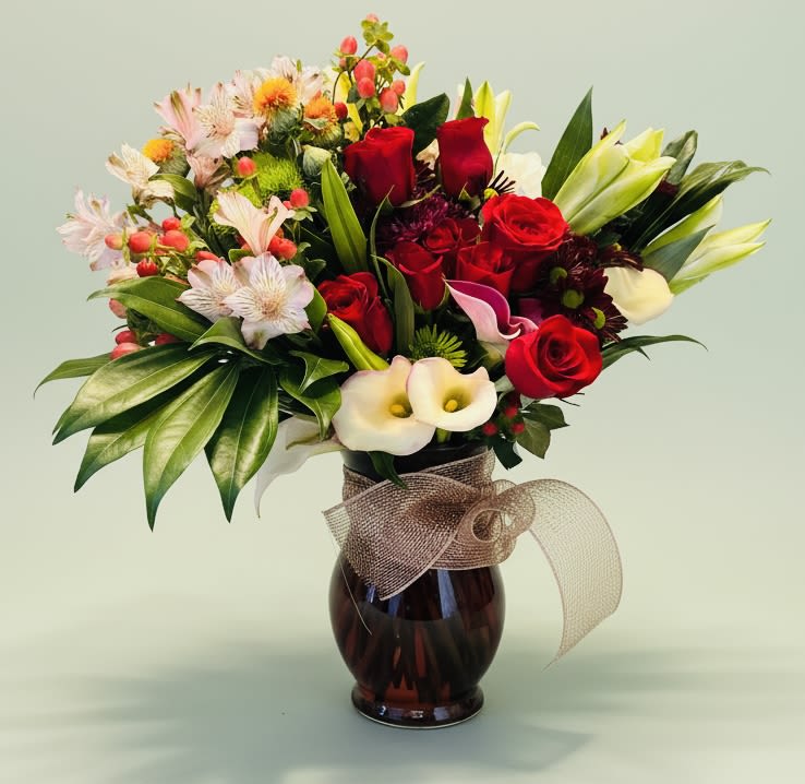 Ophelia's Opulence - A lush, rounded bouquet of red roses and white lilies anchored by soft pink and peach accents-this flower arrangement is a romantic, celebratory statement perfect as an anniversary bouquet, Valentine's Day surprise, or a thoughtful birthday gift. The visual impact of rich crimson focal blooms set against pale lilies and airy pink fillers creates an elegant contrast that will make the person you're thinking of feel truly seen and cherished. It also carries the warmth and formality to suit Thanksgiving, Christmas, or a refined sympathy or thank-you gesture.  The mix includes roses, lilies (including calla-style shapes and lily buds), alstroemeria, carnations, chrysanthemums and other delicate fillers, all layered with glossy green foliage for depth. Arranged tight and full in a keepsake glass vase and finished with a ribbon, the composition balances classic romance with a subtle rustic charm-great for housewarmings, retirement celebrations, weddings, or as a boss's day or Mother's Day treat. Size and delivery details are available at checkout; to keep blooms fresh, place the vase in a cool spot away from direct sun and change the water every two days.