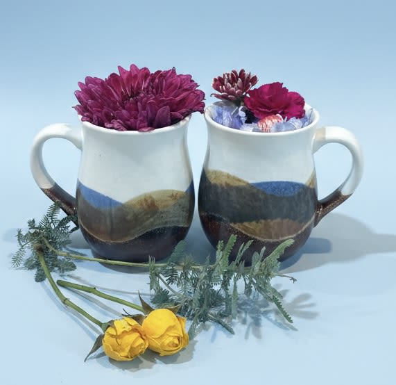 Pair of pottery mugs with wildflowers - Two cheerful ceramic mugs filled with wildflower-style blooms make a delightfully informal gift for birthdays, thank-yous, or a just-because surprise. One mug overflows with deep pink chrysanthemums while the other holds a soft mix of pale blue and pink flowers; two sunny yellow rosebuds and loose greenery sit ready to tuck in or rearrange, so the recipient immediately feels seen and celebrated. This pairing reads as thoughtful and personal-the kind of small bouquet that brightens a kitchen windowsill, office desk, or bedside table.  Each arrangement arrives in its own pottery mug, so the flowers double as lasting keepsakes the recipient can use again and again. Colors include playful blues, warm yellows, soft pinks, and clean white accents for a balanced, spring-ready palette that also suits winter and holiday gifting when paired with seasonal wrapping. Perfect for Mother's Day, Father's Day, weddings, Valentine's Day, or congratulating a coworker on Boss's Day, these mug bouquets are low-fuss: simple care is watering the floral foam or replacing water as needed and trimming stems for freshness. Order them together for a matched set that feels intimate and charming.