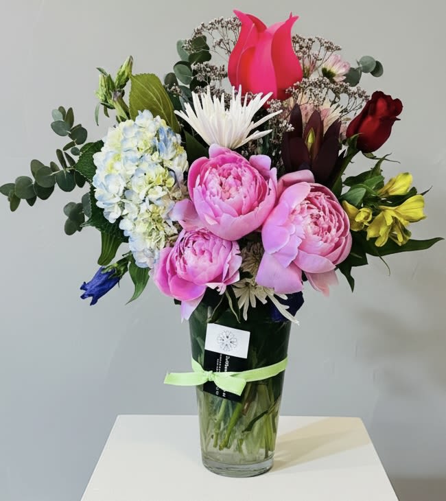 Peony Envy - Large, lush pink peonies take center stage in this romantic flower arrangement, making it an unforgettable choice for a birthday bouquet, anniversary gift, or just-because surprise. The design blends peonies with a pale blue-and-white hydrangea cluster, a single red rose, a bright pink tulip and cheerful white daisy-like blooms to create a vibrant, textured statement that reads as both joyful and deeply affectionate - perfect for Mother's Day, Valentine's Day, or a spring wedding accent.  Carefully arranged in a clear glass vase and tied with a green ribbon, the mix of rounded, layered petals and airy filler gives the bouquet a full, garden-fresh look that feels intentionally effortless. Colors range from soft pinks and whites to pops of red and pale blue, with green foliage for contrast; hard goods include floral wrap and a product tag so it arrives gift-ready. The vase size and styling make it equally at-home on a dining table, desk, or entry console.  This is the kind of arrangement that makes the person you're thinking of feel truly seen - whether you're saying thank you, congratulations, I'm sorry, or simply thinking of you. Order with standard care instructions included; fresh-cut stems benefit from fresh water every two days and a clean vase to keep blooms looking their best through spring and summer celebrations and beyond.