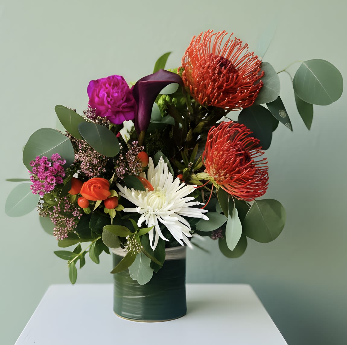 Pimpin' Protea Bouquet - Bold pincushion protea takes center stage in this lively flower arrangement, making it a striking birthday bouquet or "just because" gift that feels confidently modern. The Pimpin' Protea Bouquet blends protea, a single white chrysanthemum and deep purple calla lily with smaller purple and orange accents to create a dramatic, joyful color story-perfect for saying "thinking of you," "thank you," or congratulations for graduation and retirement.  Textural eucalyptus foliage fans around the blooms to give the design an airy, relaxed silhouette, while the dark green ceramic vase provides a polished, contemporary base you can display without fuss. Colors range from vivid red and deep purple to crisp white and warm orange, offering high contrast that reads beautifully in photos or on a tabletop. Sized to make a statement without overwhelming a room, this arrangement arrives ready to gift and enjoy; refresh water every few days and trim stems if desired to help the bouquet stay lively.
