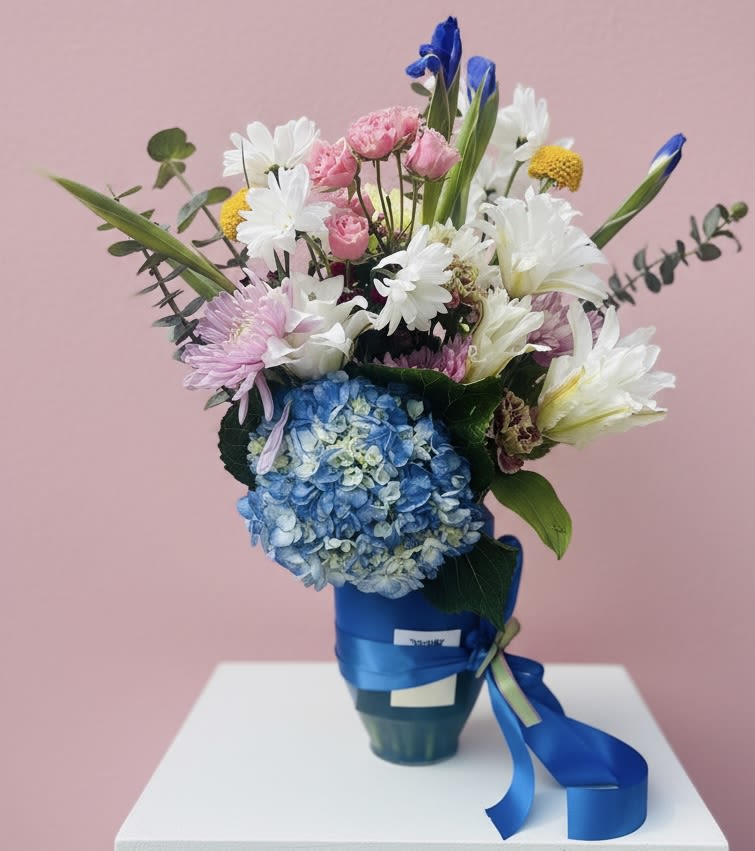 Pretty in Pastels - A bright cluster of blue hydrangea anchors this pastel bouquet, giving the arrangement a soft, cloudlike base that makes the pink roses and white lilies pop-perfect for a birthday bouquet, a thoughtful "thinking of you" gift, or a cheerful Mother's Day surprise. The mix of white daisies, blue-tipped accents, and fresh greenery creates a garden-fresh, uplifting look that will make the recipient feel genuinely seen and celebrated, whether you're saying thank you, welcoming a new baby, or marking an anniversary.  Arranged upright in a glossy ceramic vase and tied with a satin ribbon, the design balances full-bodied blooms with airy spacing and long stems for a lively, elegant silhouette. Expect hydrangeas, lilies, roses, daisies, and seasonal accents in pink, white, yellow, blue, and green; the bright blue wrap and matching ribbon finish make it gift-ready straight from the shop. Sized to sit beautifully on a mantel, bedside table, or reception desk, this pastel mix also suits spring and summer events and is appropriate for more reflective moments like sympathy or a memorial when a softer palette is desired-simple care: fresh water, a cool spot away from direct sun, and occasional stem trims to keep blooms looking their best.
