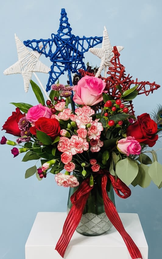 Red, White and Blue Stars Bloom - Bright, playful and distinctly festive, this red, white and blue flower arrangement is a cheerful choice for a birthday bouquet, summer celebration, or a spontaneous "just because" surprise. Red and pink roses take center stage alongside clusters of pink-and-white carnations, while decorative woven star picks in blue, white and red rise above the blooms to lend a patriotic, celebratory flair perfect for Fourth of July or Memorial Day moments.  Set in a clear glass vase with a wide red ribbon tied around the stem, the mix includes small berry accents that add texture and a touch of whimsy. The compact, vase-ready design makes it easy to place on a table or desk-an eye-catching gift that reads as both thoughtful and festive.  Send it to say thank you, congrats, or to brighten someone's summer day; the recipient will feel genuinely celebrated by the bold colors and playful stars. Vase-arranged for convenience and durability, this bouquet arrives ready to display-simply refresh the water as needed to keep blooms at their best.