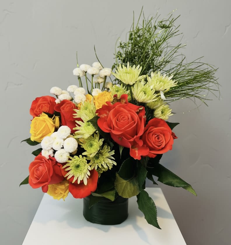 Rosy Mums - Bright clusters of red and yellow roses meet pale green daisy mums in a compact, rounded arrangement that feels both joyful and intimate - a perfect anniversary bouquet or a "just because" surprise for the person who lights up your day. Small white button chrysanthemums and airy, grass-like accents add soft texture and lift, so the recipient will notice the cheerful color pop from every angle and feel genuinely celebrated.  Arranged in a dark vase that grounds the vivid palette, this design pairs warm reds, sunny yellows, soft peaches and pink hints with crisp white and green for a lively, balanced look. The compact size makes it ideal for birthdays, Mother's Day, Valentine's Day, or romantic moments when you want a polished, everyday bouquet that still reads as special. Care is simple: fresh water in the vase and regular trimming keep blooms bright; choose same-day delivery for the most vibrant presentation.