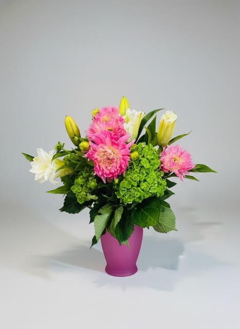 Spring Fling - A pop of joyful color that says "thinking of you" with the energy of spring-this Spring Fling flower arrangement is a bright bouquet for birthdays, housewarmings, new babies, or just-because moments. The arrangement's vivid mix of white lilies, soft pink daisy-like blooms, bold dahlias, and a lusciously green hydrangea cluster creates an uplifting contrast that reads fresh and celebratory whether you're marking a graduation, saying thank you, or sending a note of love and romance.  Arranged in a magenta rounded vase, the composition balances open, airy lilies with the dense texture of hydrangea and the cheerful charm of pink petals, finished with glossy green foliage for dimension. The compact, contemporary vase makes it easy to place on a table or counter as a centerpiece or accent; colors-green, yellow, white, and pink-work together to brighten any room. Ideal for spring and summer occasions yet perfectly suited for everyday surprises, this bouquet delivers an immediate sense of care and delight to the person you're celebrating.