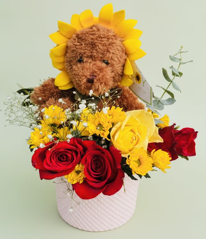sunflower Bear bouquet - A playful plush bear peeks out from a sunflower-style headpiece above a dense, cheerful bouquet-perfect as a birthday arrangement or a romantic "thinking of you" surprise. This flower arrangement blends red and yellow roses with small yellow daisy-like blooms and airy white filler to create a warm, affectionate impression that reads as both whimsical and heartfelt for occasions like love and romance, thank you, get well, or just because.  The flowers arrive nestled in a pale pink textured gift box with a gift tag, the warm brown teddy bear adding a soft, huggable touch. Colors mix bold reds and sunny yellows with touches of white and soft pink from the box, giving a lively spring or summer feel without feeling seasonal. Ideal for Mother's Day, Valentine's Day, Easter, or to say "I'm sorry," the size and composition make this arrangement easy to display on a table or bedside-no special care beyond fresh water for the blooms and a gentle spot for the plush companion.