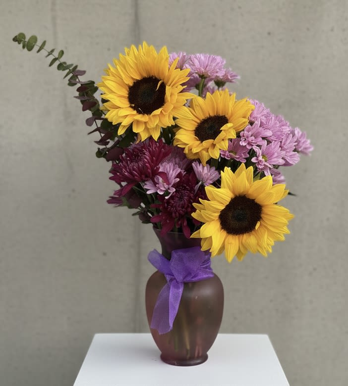 Sunflower Specialty Vase - Three bold yellow sunflowers take center stage in this sunflower bouquet for birthdays, Father's Day, or a bright just-because surprise-designed to make the person you're thinking of feel genuinely seen. The cheerful mix reads as both celebratory and comforting, a perfect pick for get well wishes, housewarmings, or to say "I'm sorry" with warmth rather than formality.  Clusters of purple and pink chrysanthemums surround the sunflowers, with a single long eucalyptus stem arcing to the left for a loose, airy silhouette. Arranged in a rounded brown vase and finished with a purple ribbon, the composition feels gift-ready and casually elegant-ideal for spring and summer tables or welcoming a new baby. Standard sizing fits most tabletops; to keep stems fresh, change the water every 2-3 days and recut stems at an angle when you refresh the vase.