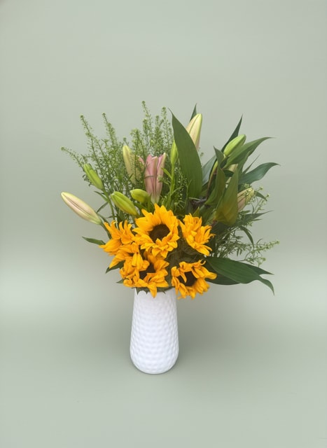Sunflowers and Lilies - Bright, sunny sunflowers take center stage in this cheerful flower arrangement, paired with pale pink and cream lily buds for a soft, elegant contrast-perfect as a summer birthday bouquet, a Father's Day surprise, or a "just because" thank-you gift. The mix of bold yellow, gentle pinks, crisp white accents, and fresh green foliage reads warm and optimistic, the kind of gift that makes the person you're thinking of feel noticed and genuinely cheered.  Clustered low in front with upright lilies rising behind, the composition is airy and natural with fine-textured greenery and long leaves that create height and movement. Presented in a clean white textured ceramic vase, the arrangement keeps a modern, fresh look that suits housewarming, wedding reception accents, thinking-of-you or get-well moments, and everyday spring or Easter celebrations. Order this sunny arrangement to send congratulations, Mother's Day wishes, or simple cheer-sizes and vase options are available to match the occasion and space.