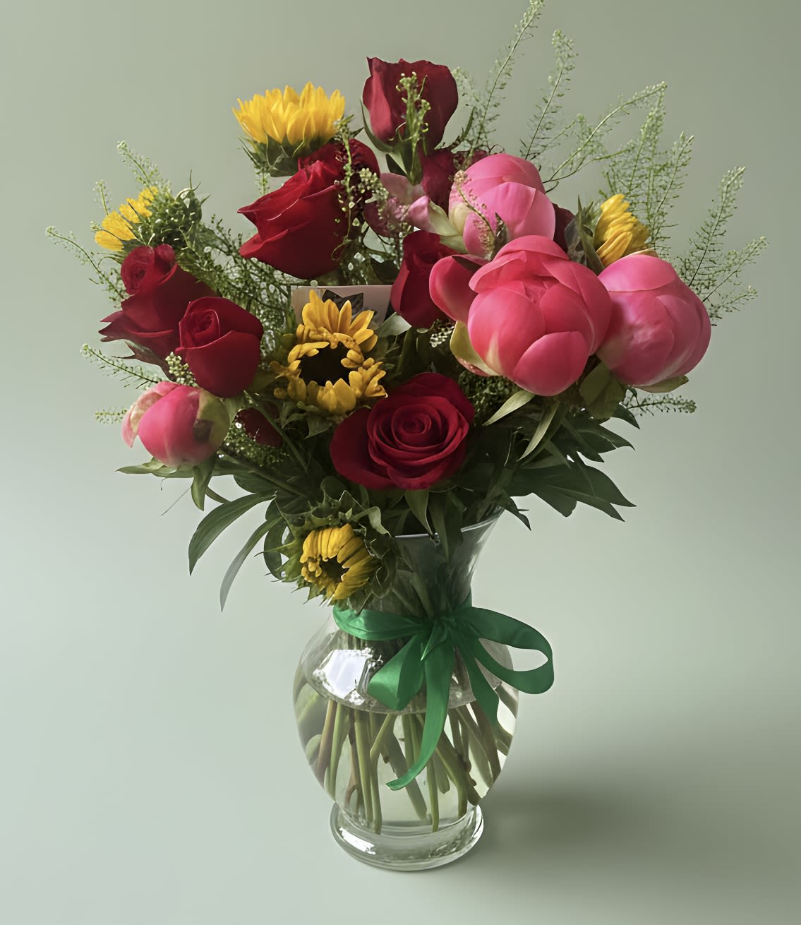 Sunny Blooms Bouquet - A vivid mix of deep red roses, bright pink peonies and sunny yellow daisy-like blooms arranged in a full, rounded bouquet-this anniversary and birthday arrangement reads as joyful and romantic at once. The saturated reds and pinks give a passionate, celebratory feel while the yellow accents lift the mood; the loose, garden-style shape and airy filler make it feel freshly gathered for the person you want to celebrate.  Hand-tied and presented in a clear glass vase with a green ribbon, the bouquet shows off each stem and keeps a natural, hand-arranged look perfect for love and romance, a new baby welcome, spring or summer celebrations, thinking-of-you moments, get well wishes, housewarmings, and thank you gestures. The colors and textures work year-round; simply place in fresh water, recut stems at an angle every few days, and enjoy the blooms as they open.