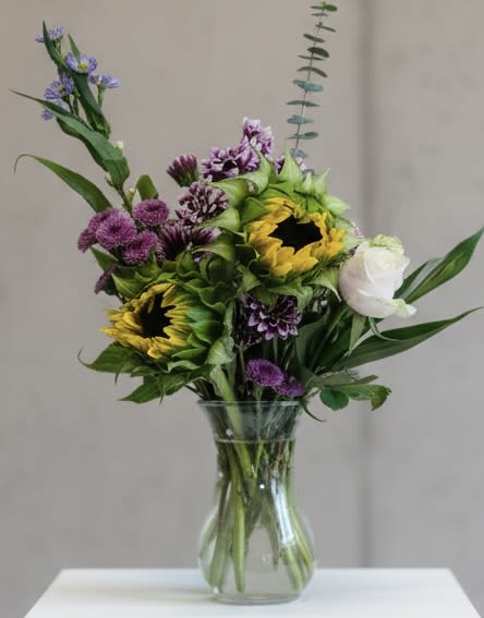 The Complimentary Couple - Two or more bold sunflowers take center stage in this cheerful mixed bouquet, their bright yellow faces set against deep purple and lavender accents for a lively, garden-fresh look-perfect as a birthday bouquet, a summer thank-you, or a thoughtful "just because" gift. The arrangement also includes a single white rose that adds a quiet, comforting note, making it appropriate for get-well wishes or a gentle sympathy and funeral tribute when a softer touch is needed. The overall effect is warm and uplifting, the kind of bouquet that makes the person you're thinking of feel seen and brightened.  Designed in a clear glass vase, the loose, upright composition uses long, arching stems and broad green foliage to create a natural, slightly rustic silhouette that works well on a dining table, desk, or hospital bedside. Flowers include sunflowers, chrysanthemums, roses, and a mix of seasonal filler blooms in purple, white, yellow, and green. Sized to feel generous without overwhelming a small space, it arrives ready to display-simple care is keeping water fresh and trimming stems-to make sending congratulations for graduation or retirement, a Mother's Day or Father's Day surprise, or a heartfelt "thinking of you" as easy as placing the vase.
