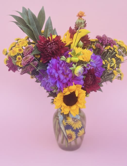 The Gypsy Sunflower - Bold sunflowers steal the show in this lively birthday bouquet, a garden-style flower arrangement made to make the person you're thinking of grin. Bright yellow sunflowers are paired with vivid purples, magentas and deep burgundy accents for a rich, layered color story that reads joyful and unexpected-perfect for Spring and Summer celebrations, a warm thank-you, or a thoughtful "just because" surprise.  The Gypsy Sunflower is arranged asymmetrically for a natural, hand-gathered look: tall foliage rises on one side while a fuller cluster of chrysanthemums and mixed seasonal blooms fills the center, giving lots of depth and texture. It's delivered in a clear glass vase with a decorative sunflower-print insert that reinforces the rustic, cheerful vibe and keeps stems neatly displayed.  Give this to someone who loves bright, energetic color-she'll feel cheerfully celebrated and seen. The vase makes it ready to display on arrival; for best life, keep blooms in fresh water and out of direct heat. Ideal for birthdays, thinking-of-you notes, or seasonal decorating throughout Spring and Summer.