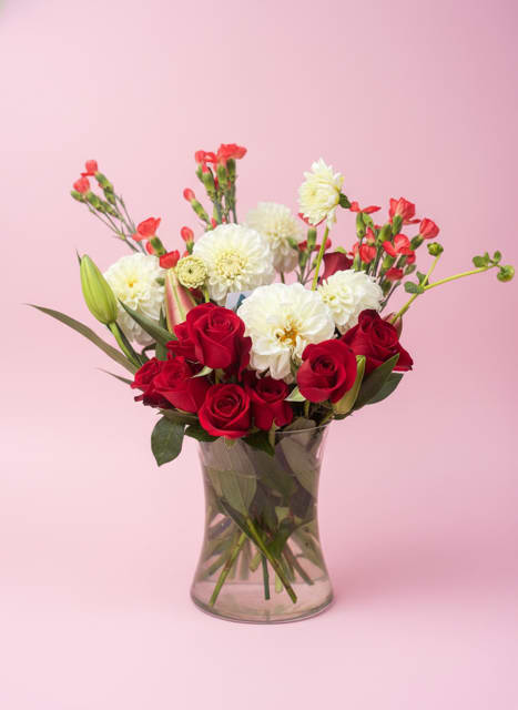 The Lovely Lush Bouquet - Velvety deep red roses paired with large white blooms and soft pink accents make this anniversary bouquet feel both romantic and effortlessly elegant - a perfect choice for love and romance, a birthday surprise, or a "thinking of you" moment. The upright, full composition invites the recipient to pause and breathe in the layered textures: rich reds grounded low, airy white stems reaching upward, and rosy tones that soften the overall look.  Hand-arranged in a clear glass vase, The Lovely Lush Bouquet features carnations, lilies, roses, dahlias, and chrysanthemums in pink, red, and white hues for a lush, mixed-flower effect. The vase creates a clean, modern profile while showcasing the stems; the design reads as both festive for weddings or Valentine's Day and simple enough for everyday thank-you or congratulations gestures.  This is the kind of bouquet that makes someone feel truly seen - intimate without being fussy. Arrives ready to display; trim stems and refresh water every few days to extend vase life.