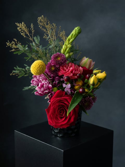The Rendevous - A single, dramatic red rose anchors this compact birthday bouquet,  (* larger ones have more roses) surrounded by vibrant pink, purple and sunlit yellow blooms to create a lively arrangement perfect for love and romance, an anniversary, or a "just because" surprise. The design reads modern and bold-the saturated colors pop against the sleek black vase-so the person you're thinking of will feel celebrated and truly seen the moment it's set down.  This flower arrangement features rounded chrysanthemum heads, tulip buds, lilies and mixed accent flowers arranged tightly with airy branching filler rising behind the main cluster, giving texture and height without losing the intimate silhouette. The low, moody black vase keeps the focus on color contrast and fits easily on a bedside table, desk, or reception counter; compact sizing makes it an ideal choice for weddings, thank-you gestures, Valentine's Day, New Year greetings, or as a thoughtful thinking-of-you gift.  Give it to someone who deserves a joyful, romantic moment-whether you're marking an anniversary, saying thank you, or simply brightening their day. Care is simple: keep water fresh, trim stems every few days, and place away from direct heat to extend blooms; this arrangement is designed to deliver immediate impact and lasting smiles.
