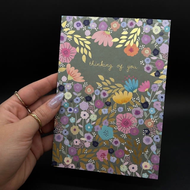 Thinking of You card - A floral-themed greeting card that looks like a pocket of sunshine-perfect as a thinking of you card for encouragement, sympathy, or a simple check-in. The dark green background makes the dense border of small illustrated flowers-purples, pinks, yellows and blues-pop like a hand-drawn bouquet, while the gold script "thinking of you" in the center delivers a warm, sincere message the recipient can keep.  Printed on cardstock with a stationery-shop, handmade feel, the design reads as both cheerful and comforting: the jewel-toned blossoms add richness while the pastel accents keep the tone gentle. Packaged with a matching envelope, this card is ready to slip into a gift, leave with flowers, or send on its own to brighten someone's day; it's the kind of note that helps a person feel seen and supported.