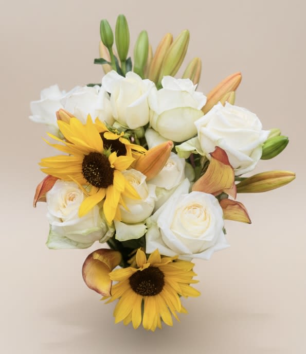 Today, Tomorrow and Yesterday Bouquet - Bright, uplifting and thoughtfully balanced, this flower arrangement-the perfect "Just Because" or Thinking of You bouquet-pairs white lilies and roses with vivid yellow sunflowers to create an cheerful, sunlit statement. Ideal for birthdays, Get Well wishes, Thank You gestures, or a Spring and Summer centerpiece, it brings the warmth of a sunny day indoors and reads as a comforting, celebratory gift for the person you're thinking of.  The mix features open white roses and lilies with several taller, unopened lily buds that add height and graceful structure, while round rose blooms soften the overall shape. Bold sunflower faces and their deep centers provide contrast and a joyful focal point against crisp white petals and fresh green foliage. Colors include white, yellow, orange highlights from the sunflower centers, and lively greens for a clean, natural palette.  Perfect for Father's Day, Mother's Day, congrats moments or to brighten a recovery room, this arrangement is designed to make someone feel seen and uplifted. Sized to work on a tabletop or entry console, it ships ready to display-trim stems, fresh water, and simple care will keep blooms looking their best so your recipient enjoys the message as long as possible.