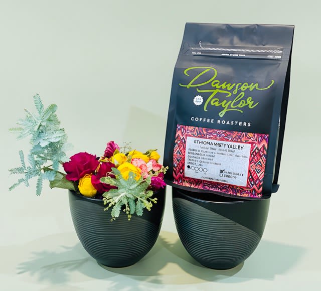 Wildflowers mini bunch and mugs w/ bag of local coffee beans - A compact burst of cheerful color that feels effortless to give, this flower arrangement for birthdays, Father's Day, or "just because" moments pairs bright wildflower-style blooms with a bag of local coffee for an uplifting gift. The mini bunch and mug-style presentation is perfect for housewarming, thank-you gestures, or to say "thinking of you"-the recipient will feel noticed and energized the moment they open it.  Yellow, magenta, pink and pale blue-green foliage mingle with carnations, roses, daisies and other meadow-like blooms, arranged in a low black plastic container that creates a modern contrast with the lively colors. The matching black container holds the compact bouquet beside an upright bag of locally roasted beans with a colorful label, so the set reads cohesive and polished on a desk, kitchen counter, or bedside table.  Ideal for occasions from anniversaries and weddings to winter holidays and Mother's Day, this gift feels both celebratory and thoughtfully useful-flowers to brighten a room and coffee to brighten a morning. The arrangement is delivered in its black container with the coffee bag included; for best enjoyment, keep flowers in fresh water and away from direct heat, and let the recipient know the coffee is locally roasted for a fresher, more personal cup.
