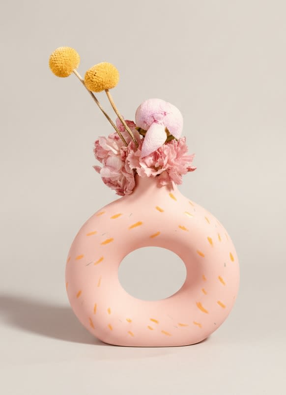 The Donut Vase ( Minimalist Design) can be delivered with a box of assorted donuts  - A pastel pink donut-shaped ceramic vase with orange speckled accents makes a cheerful, modern statement-perfect as a casual birthday bouquet, spring pick-me-up, or "thinking of you" gift. The minimalist design keeps the focus on the recipient: compact, whimsical, and bright enough to make someone feel noticed without overwhelming their space.  A small cluster of soft pink carnation-like blooms crowns the top, balanced by two tall yellow pom-pom stems and a pale white bud for contrast and height. The arrangement is intentionally light and decorative rather than a full bouquet, designed to sit on a desk, windowsill, or bedside table. The ceramic vase is the highlight-donut-shaped, pastel pink with subtle orange speckles-adding texture and keepsake value.  This is an ideal choice when you want the person you're thinking of to feel cheerfully seen; it pairs beautifully with everyday celebrations and casual gifting moments. Optionally delivered with a box of assorted donuts for an extra treat-small scale, low-maintenance, and ready to display right out of the box.
