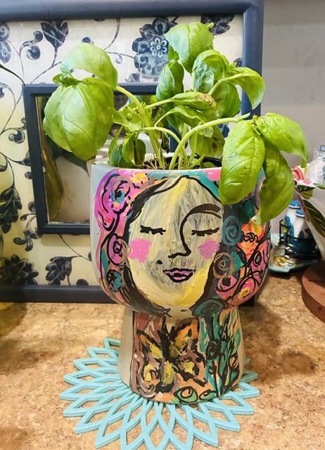 Custom painted face vace pots each one is uniquely painted - A playful, artful touch for a housewarming or casual gift-this leafy green arrangement in a hand-painted ceramic face planter brings color and personality to any room. Perfect as a housewarming gift or a cheerful "thinking of you" present, the stylized face design and bright accents make the recipient feel seen and delighted the moment it arrives.  Glossy broad green leaves nestle in a decorative ceramic planter hand-painted with pink, blue, yellow, black, and orange accents and mounted on a painted stand for added display height. The contrast between the fresh foliage and the whimsical, colorful face gives a modern, artsy look that suits living rooms, entryways, or home offices.  Ideal for someone who loves unique decor, this piece arrives ready to brighten a shelf or tabletop and makes a memorable housewarming or hostess gift. Simple care-regular bright, indirect light and occasional watering-keeps the leaves glossy and the planter the focal point of the room.
