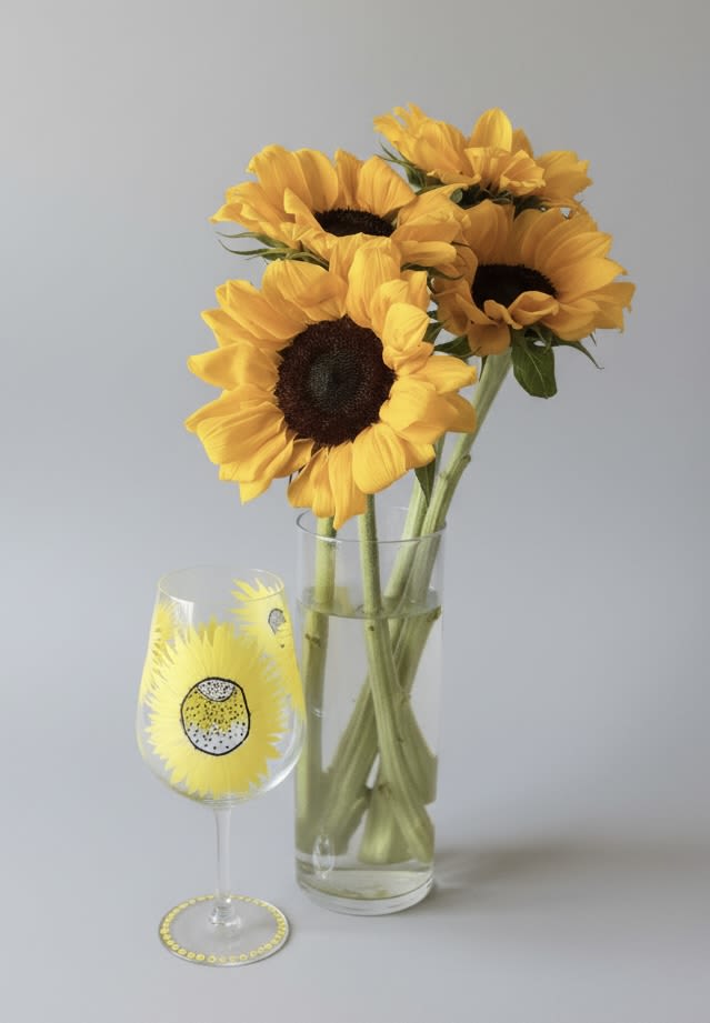 Painted Sunflower wine glass with sunflowers vase - Bold, sunlit sunflowers take center stage in this cheerful flower arrangement-perfect as a birthday bouquet, a "thinking of you" gift, or a sunny seasonal surprise. The bright yellow blooms with their dark, dramatic centers create an immediate warm, uplifting impression the recipient will notice from across the room. The presentation pairs the floral display with a painted sunflower wine glass for a playful, coordinated accent that makes the gift feel intentionally styled.  Arranged in a clear cylindrical glass vase, the full, open sunflower heads are complemented by fresh greenery and subtle white highlights that add texture and balance without competing with the blooms. The clear vase showcases clean lines and keeps the focus on the flowers' warm tones, while the matching wine glass-decorated with a sunflower motif-adds a charming keepsake the recipient can enjoy long after the stems fade. This casual, sunny combination suits everyday gifting, cheerful get-well wishes, or as a bright centerpiece for seasonal gatherings.  Choose the size that best fits your occasion; arrangements arrive ready-to-display with water and simple care instructions included so the recipient can enjoy long-lasting blooms. Whether you're celebrating a friend, brightening a kitchen, or sending a note of encouragement, this sunflower pairing delivers a genuine, sunlit smile.
