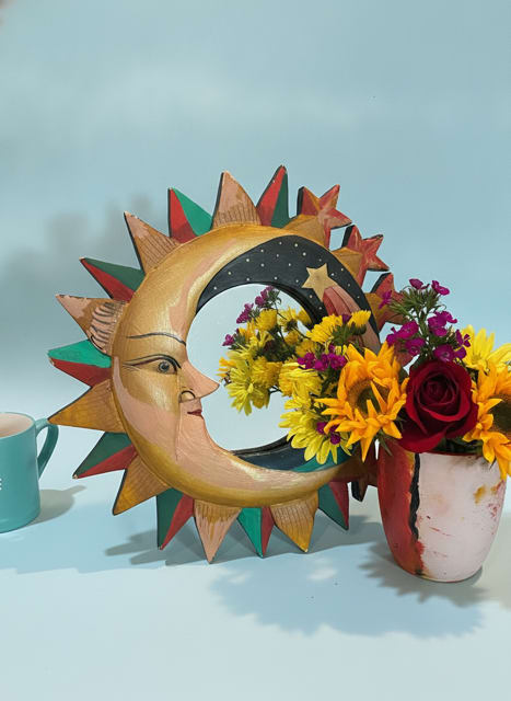 painted mirror and sunflowers bouquet bundle - Bright, folk‑art charm meets a cheerful bouquet in this painted mirror and sunflowers bouquet bundle - an ideal everyday gift or birthday bouquet for someone who loves color and whimsy. The combination of a sun‑face wall sculpture or painted mirror with a compact floral arrangement sends a playful, sunny message that will make the person you're thinking of feel noticed and celebrated. This is the kind of gift that brightens a kitchen wall and the recipient's day at once.  The arrangement pairs bright yellow daisy‑like blooms, a single deep red rose, and small magenta filler flowers tucked into a hand‑painted ceramic vase, complemented by greenery for texture and balance. Accents include folk‑art hard goods: choose the decorative sun wall sculpture or the painted sun‑and‑moon mirror with multicolored rays, both lending a handcrafted, cottage‑style vibe. The palette reads joyful - yellow, red, pink, blue and green - and the mix of roses and chrysanthemums (plus other seasonal filler) keeps the bouquet feeling lively and fresh.  Perfect for birthdays, housewarmings, or a "thinking of you" surprise, this bundle is curated to create an immediate visual lift in a casual home setting. The compact size makes it easy to place on a small table, shelf, or entryway; pair it with the ceramic mug or wooden wall art for an upgraded gift set. Standard care: change vase water regularly, trim stems at an angle, and keep away from direct heat to prolong freshness.