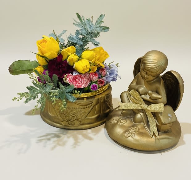 Cherub box with mini roses and wildflowers - A compact cluster of sunlit yellow roses crowned by a petite gold angel creates an immediate sense of warmth and comfort-perfect as a sympathy arrangement or a tender memorial gift. The Cherub box with mini roses and wildflowers reads like a small, ceremonial keepsake: bright yellow roses bring cheer, while pink, purple, and pale blue accents add gentle color notes that feel both lively and consoling for the person you're honoring.  Arranged in an ornate gold decorative container with a matching gold ribbon and angel figurine, this piece pairs structured charm with airy, blue-toned foliage for a soft, textured silhouette. Small wildflower accents and mixed-color sprays surround the mini roses to create visual depth in a compact footprint-an elegant tabletop size that fits mantels, bedside tables, or a reception counter without overwhelming the space.  Give this to someone who needs to feel seen-whether for a remembrance, a get-well gesture, or a quiet way to mark an anniversary-so they'll sense care and thought in every detail. The arrangement arrives ready to display in its gold container; keep blooms in fresh water and away from direct heat to extend their cheerful presence.