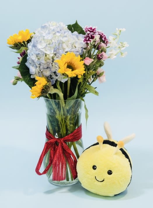 The Bumble bee bouquet - Bright sunflowers and a cuddly plush bee make this cheerful flower arrangement a joyful pick-me-up or birthday bouquet that says "thinking of you" with sunny warmth. The Bumble Bee Bouquet is ideal for everyday gifting, a cheerful thank-you, or brightening someone's recovery-she'll feel genuinely seen by the playful mix of colors and the little bee companion.  A generous cluster of pale blue‑white hydrangeas anchors the design, contrasted with vibrant yellow sunflowers and smaller pink and purple daisies and carnations for delicate pops of color. Delivered in a clear glass vase , the mixed bouquet arrives gift-ready and visually striking from every angle. The plush bee sits beside the vase to reinforce the whimsical, sunny theme.  Perfect for friends, coworkers, or anyone who could use a lift, this arrangement balances bold focal blooms with soft, textured accents to create an uplifting presence in a room. Sizes and exact counts vary by season; vase and ribbon are included, and simple care-fresh water and a cool spot-will keep the flowers looking their best through the week.