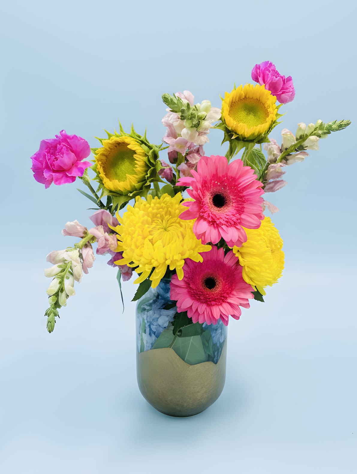 Striking Summer Bouquet - Hot pink gerbera daisies meet sunny yellow sunflowers and cheerful chrysanthemums  with pink  snap dragons in a Striking Summer Bouquet  set into soaking  powdery-blue hydrangeas flowers as the base-built to make someone feel instantly celebrated. This birthday bouquet, thank-you arrangement, or just-because gift delivers a high-energy contrast of hot pink, bright yellow, soft whites and fresh greens, with pastel snapdragon spikes adding vertical texture and a playful, airy silhouette that reads joyful from across the room.  Arranged loosely in a rounded, breathable composition, the blooms come presented in your choice of a tall clear glass vase with a muted metallic lower section, a gold-toned rounded vase, or a matte metallic vase for a modern touch. Smaller mixed-vase options are shown grouped with decorative filler stones for a casual, fresh-cut look-perfect for tabletops or office desks. The mix includes gerbera daisies, sunflowers, snapdragons, chrysanthemums, dianthus and bright filler flowers, creating a lively, long-lasting display.  Choose this bouquet for birthdays, springtime celebrations, a cheerful thank-you, or to brighten someone's everyday space; the recipient will feel genuinely seen and uplifted. Vases and visible filler vary by size selection-refer to the product options when ordering for exact vase style and arrangement size. Care: keep water fresh and trim stems every few days to prolong blooms.