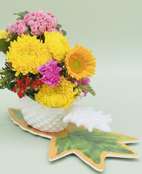 Pretty as a pineapple pot - A bold cluster of sunlit color greets the recipient the moment they see this cheerful flower arrangement-perfect as a birthday bouquet or an everyday celebration to brighten someone's day. The composition reads playful and uplifting, ideal for sending congratulations, thank-you wishes, or a spontaneous "thinking of you" surprise that makes her feel truly noticed.  Tightly gathered yellow chrysanthemums and a standout orange-yellow sunflower create the arrangement's sunny core, while pink and white carnations and mixed bouquet accents add soft contrast and texture. Red touches punctuate the palette for a lively pop against the softer hues. All blooms are arranged in a white textured ceramic vase set on a decorative pineapple-shaped base, giving the piece a charming, gift-ready presentation that looks great on a kitchen table, desk, or bedside.  Designed to arrive full and vibrant, this compact pot-style arrangement is easy to place and enjoy-no complicated setup required. To keep the flowers looking fresh, place in a cool spot away from direct heat and refresh the water every couple of days; for same-day gifting or scheduled delivery, choose your preferred delivery date at checkout.