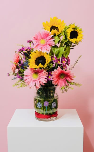 Valentina painted Daisy and sunflower vase - Bright yellow sunflowers and pink gerbera daisies take center stage in this cheerful flower arrangement, the perfect birthday bouquet or thank-you gift to brighten someone's day. The tall, open composition reads casual and upbeat-ideal for springtime celebrations or any moment you want the person you're thinking of to feel seen and joyful. (*It has been hand-painted by an eleven year old artist) It helps to further youth art development and growth.  Mixed with small purple filler blooms for contrast, the painted-daisy look brings bold color and a playful texture that reads fresh from across the room. Stems are gathered in a clear glass vase tied with a simple red ribbon for a ready-to-gift presentation; the arrangement stacks color without feeling fussy. Size and stem length create an airy, upright profile that sits nicely on a table or desk-no special care beyond fresh water and a cool spot will keep these gerbera daisies perky for days.