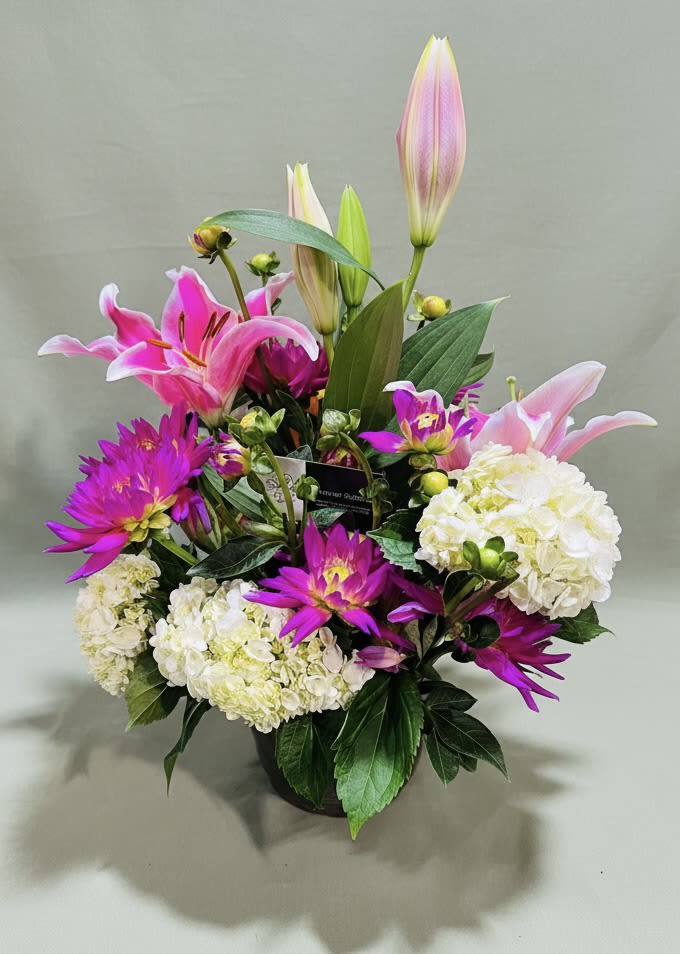 Eternity Garden Bouquet - A lush mix of bright pink lilies, vivid magenta daisy-like chrysanthemums, and soft white hydrangea clusters makes this flower arrangement an uplifting "just because" gift that reads like a promise of joy. As a bouquet for birthdays, anniversaries, thank-you moments, or a tender sympathy gesture, the Eternity Garden bouquet delivers a feminine, celebratory presence-perfect for Mother's Day, Valentine's Day, Spring celebrations, or to mark special cultural holidays like Rosh Hashanah or Nowruz without feeling seasonal.  The compact ceramic vase holds a dense, layered composition: rounded white hydrangeas form a creamy base while tall pink lilies and pale pink lily buds rise above, adding height, movement, and fragrance. Vivid purple chrysanthemum-like blooms punctuate the arrangement for lively contrast, and a clean floral wrap keeps the presentation polished for immediate gifting. Sized to sit comfortably on a side table or desk, it's arranged for strong visual impact and easy display.  This bouquet is made to make the person you're thinking of feel truly seen-whether you're saying thank you, celebrating an anniversary, sending birthday wishes, or offering comfort. Choose it when you want a polished, ready-to-present arrangement that balances lasting structure with fresh-cut scent; care is simple-trim stems and refresh water to extend bloom life.