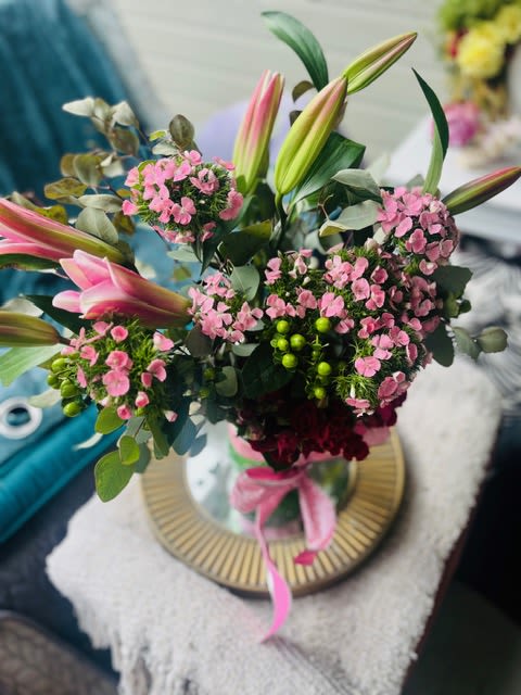 Pretty In pink Lilies bouquet - Soft pink lilies unfurl above a cloud of petite pink dianthus and carnations, making this flower arrangement an elegant choice for anniversaries, birthdays, or a tender "thinking of you" moment. Perfect as a Mother's Day or Father's Day surprise, or to welcome a new baby, the arrangement reads as warm and celebratory-she'll feel genuinely seen when these fragrant lilies and blush blooms arrive.  Clusters of soft pink petals contrast with deep green foliage and bright green berry accents for a lively, garden-fresh look. The bouquet is arranged loose and airy for natural movement, with taller lily buds giving vertical drama and rounded pockets of dianthus and carnations adding texture. Presented in a clear glass vase tied with a pink ribbon and placed on a round decorative tray, it arrives gift-ready for housewarmings, get-well wishes, sympathy, or simple summertime cheer.  This versatile design suits spring and summer moments yet remains thoughtful year-round for thank-you gestures, romantic occasions, or moments of comfort. Standard care: keep stems in fresh water, recut stems at an angle if needed, and display away from direct heat to extend bloom life.