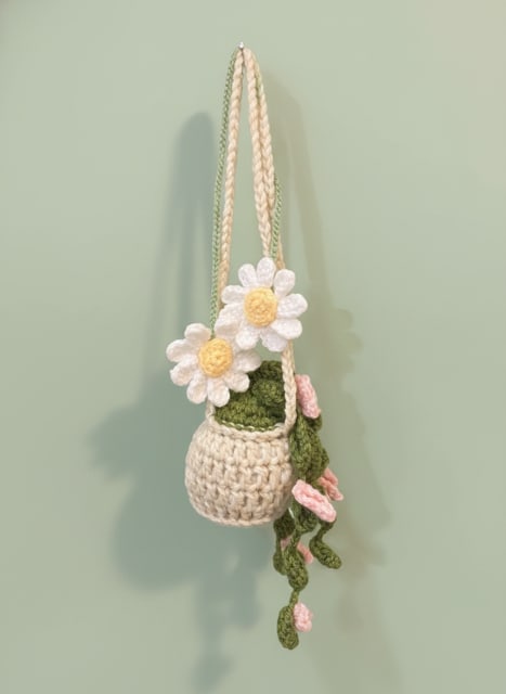 hanging crocheted flowering plant - A sweet, handmade touch that makes any room feel cared-for - this hanging crocheted flowering plant is a charming decorative accent perfect for a birthday, housewarming, anniversary, or "just because" gift. The piece reads as an everyday accent with a whimsical, tactile presence that will make the person you're thinking of feel seen and remembered whether you're marking love and romance, saying thank you, or celebrating Best Friends Day.  The small cream-colored basket is crocheted with soft yarn and hangs from a braided suspension cord, creating a rounded, cozy silhouette. Two white daisy-like blooms with sunny yellow centers sit at the front while trailing green vines and tiny pink blossoms cascade down one side; a few succulent-style stitched leaves add textural contrast. Colors are gentle - white, pink, green, touches of yellow and neutral cream - so it fits easily into spring decor or year-round display.  Ideal for gifting to someone who loves low-maintenance, handmade décor, this piece brightens a kitchen nook, entryway, or balcony without any watering or care. Its compact size makes it an easy add-on for romantic gestures, thank-you surprises, thinking-of-you moments, or a thoughtful housewarming present.