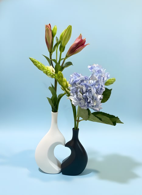 Part of My Heart Bouquet - Soft pink-and-green lily buds stand tall beside a cloud of pale blue hydrangea in a modern two-vase pairing that feels like a quiet breath of spring. This flower arrangement for everyday gifting or a thoughtful housewarming blends lilies and hydrangeas with blue, white, pink, and green tones to create a calm, airy impression that makes the recipient feel genuinely seen.  Each sculptural ceramic vase holds a distinct element: one vase with the elongated lily stems and the other with a rounded cluster of hydrangea and broad leaves, the contrasting white and dark ceramics giving the composition balanced, contemporary style. The design reads well on a mantel, entry table, or bedside-no fussy extras, just fresh texture, soft color, and clean lines.  Perfect for celebrating a new home, saying you're thinking of them, or brightening a spring morning, this arrangement arrives ready to display in its ceramic vessels. Keep flowers in fresh water, trim stems slightly if needed, and rotate out of direct heat to extend their bloom-an easy, elegant gift that comforts and delights.