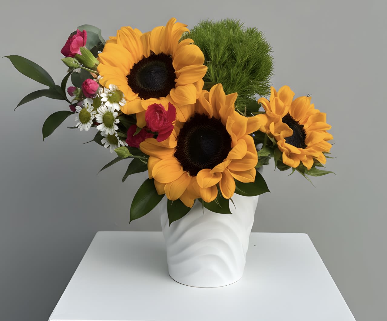 Whimsical Sunflowers - Three  or so bold golden sunflowers take center stage in this cheerful flower arrangement, designed to make someone feel genuinely seen. Perfect as a birthday bouquet, a summer housewarming gift, or a just-because surprise, the mix of sunflowers with bright pink accents and small white daisy-like blooms creates a playful, upbeat look that reads sunny and sincere-an especially warm choice for Father's Day or to celebrate retirement.  The loose, asymmetrical composition pairs three large sunflowers with chrysanthemums, mixed seasonal blooms, and green foliage for texture and movement. Pops of orange and red mingle with soft whites and pinks around the dark sunflower centers, all arranged in a clean white ceramic vase with a subtle wavy texture that fits modern and cottage styles alike. Sized to sit on a table or mantel, this arrangement brings vibrant color and layered texture without overcrowding the space.  Send this as a thank-you, housewarming, Mother's Day or Boss's Day gesture, or simply to brighten a friend's day; the cheerful palette and easy-care stems keep the mood light. For best longevity, refresh water every two days and keep away from direct heat-this is the kind of bouquet that keeps delivering smiles throughout spring and summer and works year-round whenever you want to lift someone's spirits.