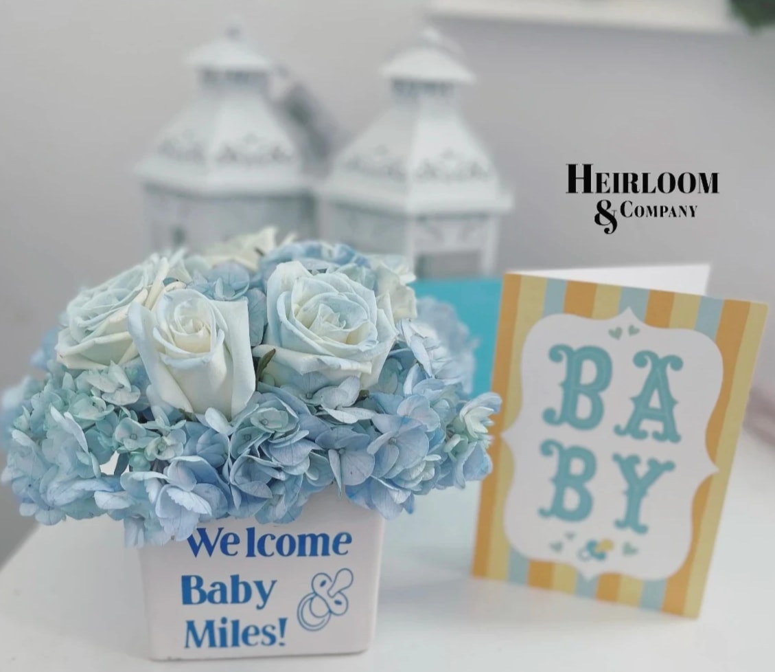 Blue and white flower arrangement in a white container with a baby card