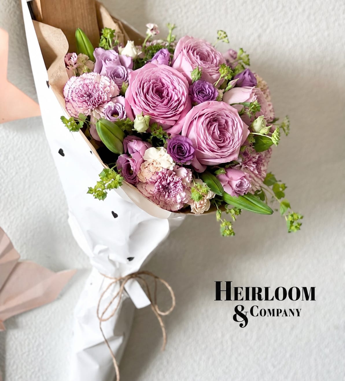 Lavender Love Letter Handheld Garden Rose Bouquet  - A romantic lavender and blush bouquet designed with premium garden roses, soft purple lisianthus, textured carnations, delicate greenery, and seasonal accent blooms. Wrapped in Heirloom &amp; Company’s signature white heart paper and finished with a natural twine bow, this bouquet is perfect for birthdays, anniversaries, sympathy gestures, thank-you gifts, or anyone who loves soft feminine florals with a luxury garden feel.  Short listing description:  A soft lavender and blush garden-style bouquet featuring premium roses, lisianthus, carnations, and seasonal accents, wrapped in our signature Heirloom &amp; Company paper.