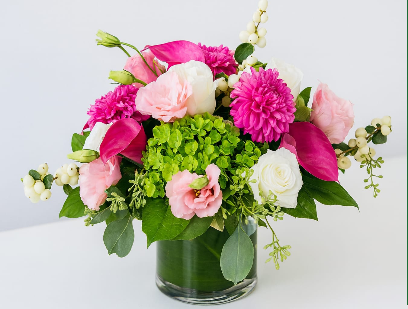 Blush &amp; Fuchsia Harmony - Bright, bold, and beautifully balanced—this arrangement is a celebration of modern elegance. Featuring radiant fuchsia dahlias, delicate pink lisianthus, creamy white roses, and sleek magenta calla lilies, it bursts with both charm and sophistication. Accented with lush green hydrangea and seasonal textures, this piece is designed in a clear glass cylinder vase lined with fresh greenery for a clean, polished look.  Perfect for brightening a dining table, elevating an office, or gifting someone special, Blush &amp; Fuchsia Harmony brings a touch of joy and style to any setting.