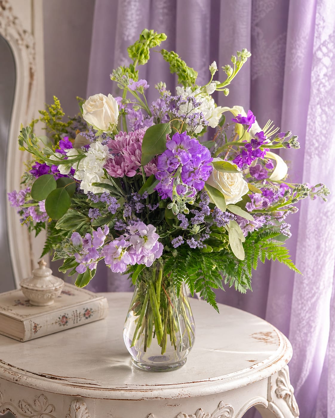 Lavender dreams - Soft lavender and creamy white blooms drift together in a garden-fresh composition that feels both romantic and effortlessly modern-perfect as a birthday bouquet, anniversary arrangement, or spring gift. Lavender Dreams is designed to make the person you're thinking of feel gently celebrated: the pastel purple notes and touches of pink read as feminine and thoughtful, while the airy silhouette adds an elegant, unpretentious charm.  The arrangement combines cream roses, pale purple stock, chrysanthemums, and clustered purple accent flowers arranged with branching, tall stems for a layered, loose look. Hues of purple, white, green, and soft pink mingle across a full, slightly romantic vase arrangement; a clear glass vase keeps the presentation light and lets the blooms take center stage. The tall, branching accents give height and movement, creating a graceful display that suits a mantel, dining table, or bedside.  Ideal for birthdays, anniversaries, or simply sending a springtime hello, this bouquet reads as both intimate and celebratory-the kind of gift that helps someone feel truly seen. Delivered in a clear glass vase, it arrives ready to enjoy; change the water every few days, trim stems at an angle, and keep away from direct heat to help the arrangement last.   If any design elements are of major importance to your order, please include them in the florist instructions at checkout or contact us to ensure availability.