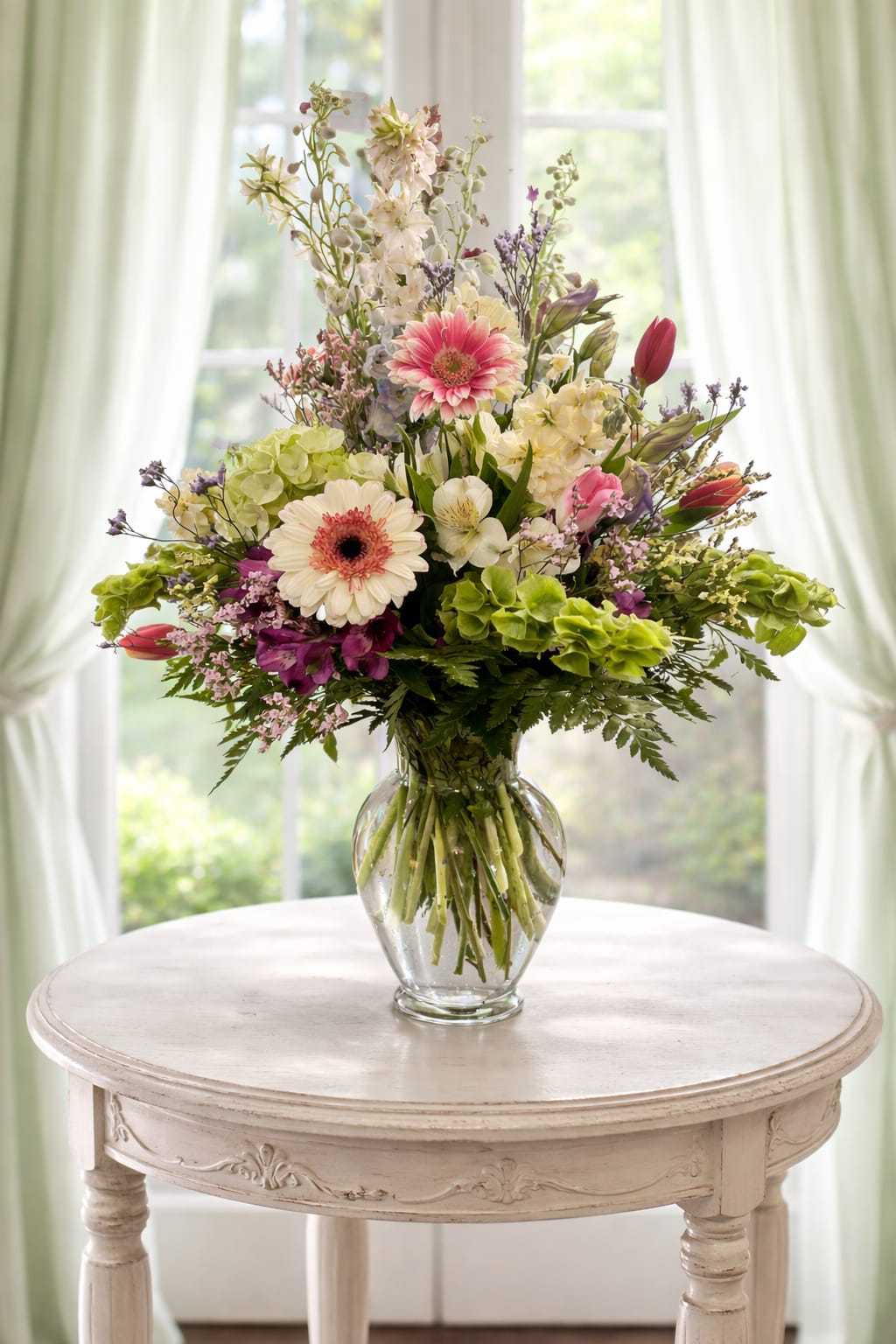 Spring bloom  - A tall, airy Spring bloom arrangement makes an elegant statement centerpiece or romantic gift for a spring celebration, wedding, or simply to brighten someone's day. The loose, garden-style silhouette is built with branching stems and layered blooms so the person you're thinking of will feel delighted by its fresh, lightly formal presence on a dining table or entry console.  White gerbera daisies and pale hydrangea clusters set a soft, luminous base while pink tulips and purple accent flowers add gentle contrasts across the composition. Alstroemeria and other seasonal filler bring delicate texture and movement; the bouquet is presented in a clear glass vase that lets the stems show through and keeps the overall look bright and breezy. Its height and fullness read as a statement without feeling heavy-perfect for a centerpiece or a meaningful gift.  Ideal for mothers, brides, or anyone who loves pastel spring color, this arrangement communicates thoughtfulness and celebration. Keep blooms in fresh water, recut stems periodically, and place away from direct heat to prolong vase life; order in time for the occasion to ensure it arrives looking its freshest.   If any design elements are of major importance to your order, please include them in the florist instructions at checkout or contact us to ensure availability.