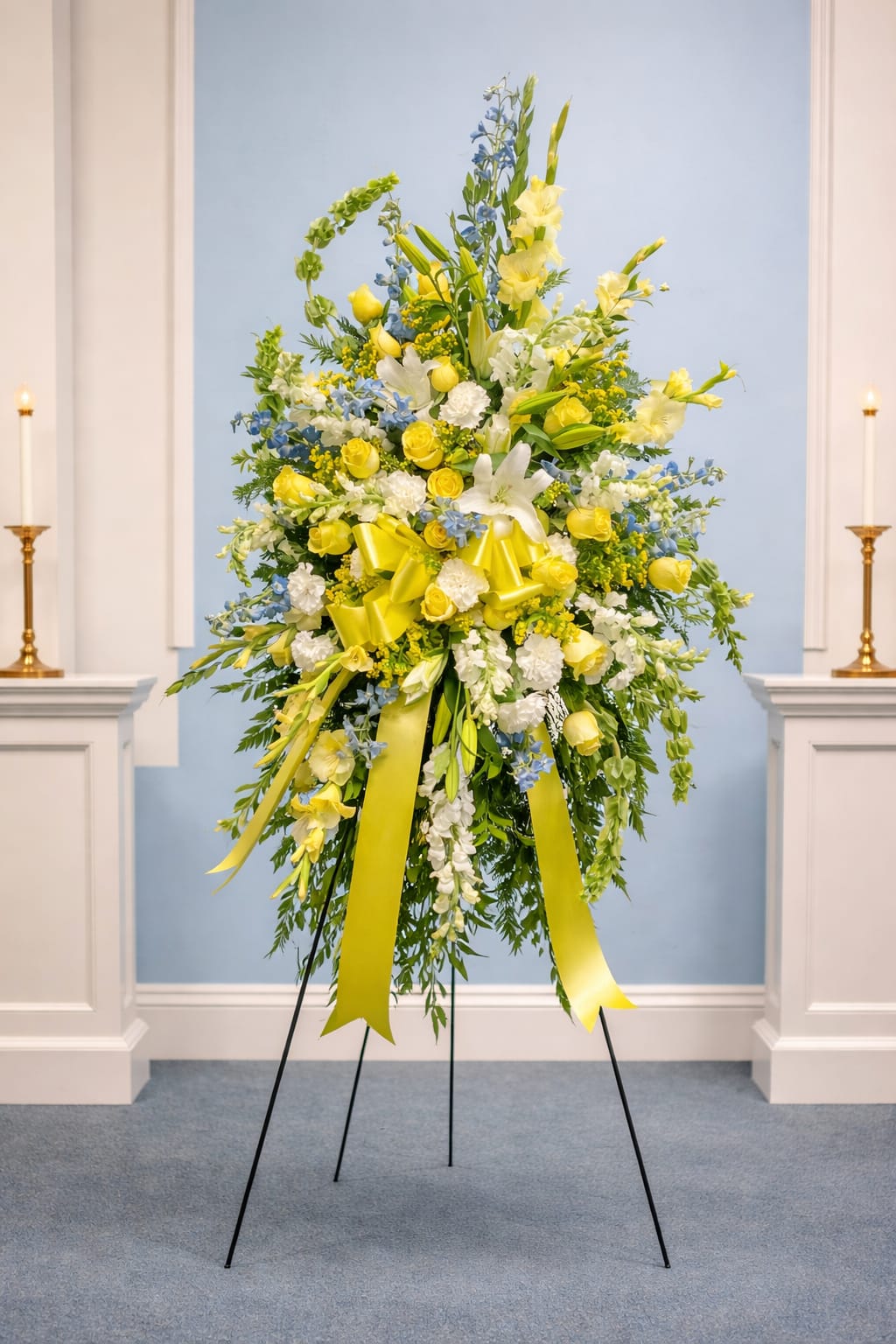 Sunrise memories - A radiant standing spray that greets a room like the first light of morning - perfect as a celebratory or sympathy floral for a funeral, memorial service, or other significant event. This tall standing spray arrangement places the flower arrangement and occasion front-and-center: the vertical silhouette and ceremonial yellow ribbon feel intentional and uplifting, the kind of display that helps the person you're honoring feel seen and remembered.  Big white and yellow lilies and roses form the focal points, surrounded by clusters of carnations, snapdragons and smaller filler blooms in blue and green tones for contrast and depth. The composition is dense and layered with airy spikes that add height and movement; bright yellow ribbon streamers are woven through the center and hang gracefully to emphasize the piece's vertical line. Arranged on a sturdy black metal easel, this formal spray reads as both elegant and hopeful.  Designed to stand at attention beside a casket, podium, or memorial table, this arrangement sends a message of warmth and respect for the person you're honoring. Care is handled by the shop - the spray arrives ready to display on its easel; for questions about delivery timing or venue setup, please contact us so we can ensure it's placed exactly where it will have the greatest meaning.   If any design elements are of major importance to your order, please include them in the florist instructions at checkout or contact us to ensure availability.
