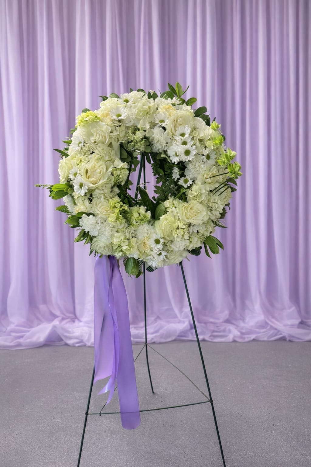 Circle of love - A large circular wreath of white roses, pale daisies, and soft hydrangeas creates a serene, ceremonial statement-perfect as a sympathy arrangement or a respectful wedding memorial. The elevated presentation on a sturdy metal easel and the trailing lavender ribbon give the piece a formal, tender presence that reads both comforting and gently romantic for the person you're honoring.  This arrangement features clustered white roses, daisy-like blooms, and full hydrangea clusters woven with fresh green foliage in a round wreath form. Mounted on a black metal easel stand and finished with a long purple ribbon draped from the lower left, its pale white, green, and purple palette is understated yet elegant. The size and wreath shape make it visible from a distance, creating a graceful focal point at services or ceremonial spaces.  Choose this wreath when you want the recipient to feel deeply respected and remembered-the design conveys sympathy, reverence, or celebratory remembrance with quiet beauty. Easy to display on the included easel; no additional assembly required.  If any design elements are of major importance to your order, please include them in the florist instructions at checkout or contact us to ensure availability.