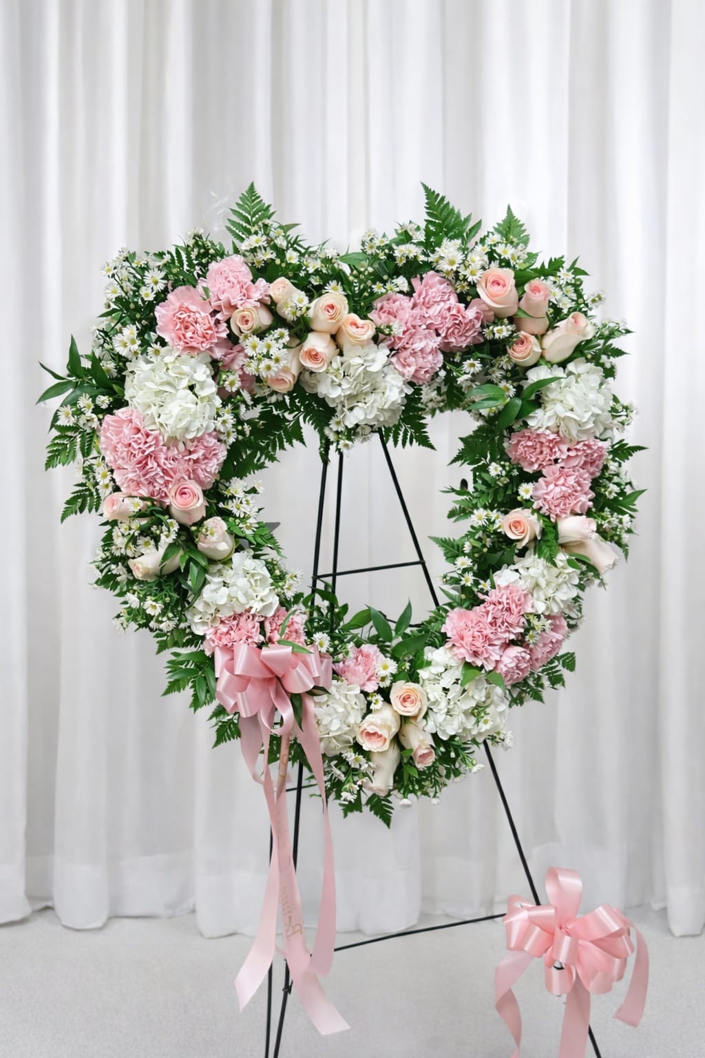 Heavenly - A tender heart of pale pink and white-this heart-shaped floral arrangement on a black metal easel is a sympathetic and graceful choice for a wedding tribute or a memorial display. The shape and soft palette communicate love and remembrance, making it a thoughtful way to honor someone special or to offer comfort to grieving family and friends.  Clusters of blush roses, pale pink carnations, and full white hydrangeas are arranged with small white daisy-style filler flowers and fern-like greenery for a lush, symmetrical presentation. Pink ribbon bows accent the design and the metal easel gives it a formal, ceremony-ready posture that will hold its shape throughout the service or event.  Ideal for funerals, memorial services, or as a heartfelt wedding tribute, this arrangement sends a message of gentle sympathy and lasting affection to the recipient. The stand-ready format makes delivery and placement easy-no additional assembly required-while simple care (keeping flowers cool and sheltered from direct sun) will help maintain its fresh appearance through the occasion.   If any design elements are of major importance to your order, please include them in the florist instructions at checkout or contact us to ensure availability.