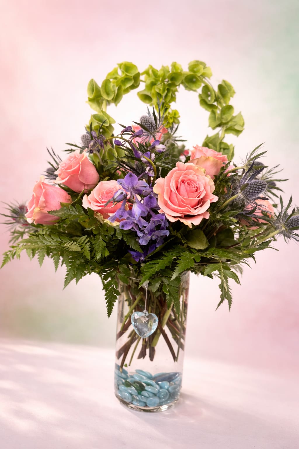 Romance - Soft pink roses take center stage in this romantic everyday bouquet, paired with small purple accent blooms and airy green foliage to create a gentle, cheerful mood perfect for a birthday, thank-you, or love-and-romance gift. The combination of warm pink and cool purple tones gives the arrangement a slightly whimsical, tender feel that makes the person you're thinking of feel truly seen and celebrated.  Arranged in a clear cylindrical glass vase, the stems sit over a bed of blue decorative glass stones that add a cool, sparkling contrast and keep the design feeling fresh. A dangling heart ornament completes the presentation, turning the piece into a ready-made keepsake. The vase size is ideal for tabletops or bedside tables; soft textures from the roses and the delicate sprays of purple provide visual depth and a light, pleasing scent.  This thoughtful bouquet suits many occasions-from cheering someone up to marking a small anniversary or saying thanks. No special care is needed beyond fresh water and trimming stems every few days to extend bloom life; the decorative stones and ornament make it an effortless, gift-ready choice for the person you want to make smile.   If any design elements are of major importance to your order, please include them in the florist instructions at checkout or contact us to ensure availability.